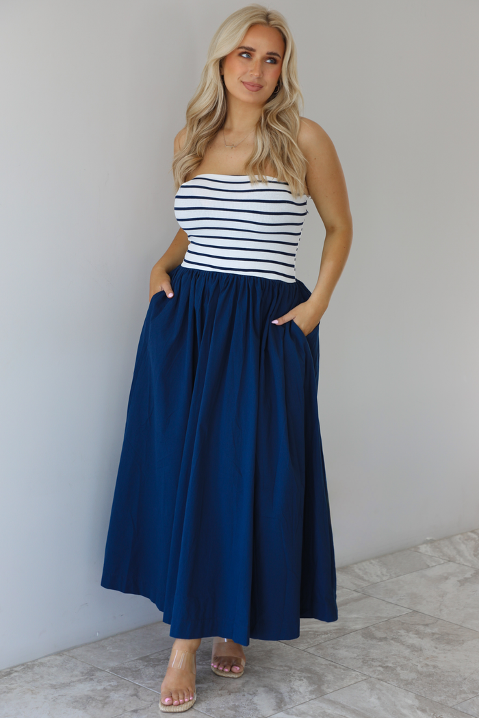 Woman wearing a maxi dress featuring a navy and white striped bodice and navy, maxi length skirt. The photo is taken indoors against a white wall with white marble flooring. 
