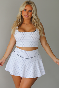 A woman modeling a white two-piece lounge set with small black polka dots, featuring a scoop-neck crop top and a matching flared skirt, standing indoors against a white wall.