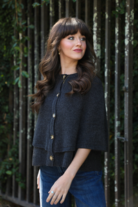 Close up of a woman model outdoors wearing a charcoal grey colored cape cardigan with functional button closures in the front and dark wash denim. 
