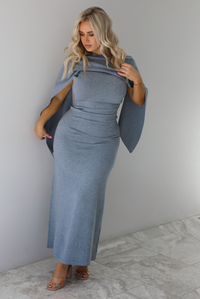 Woman wearing a grey maxi length knit dress that has a high neckline with an attached shawl detail that covers the upper half of the chest and flows on either shoulder in the back. The dress is fitted with a slightly flowy lower hem. The dress is paired with clear strapped nude heels. The photo is taken indoors against a white wall with white marble flooring. 