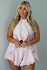 A woman modeling a light pink two-piece outfit consisting of a sleeveless high-neck ruffled top and matching ruffled shorts, standing indoors against a white wall.
