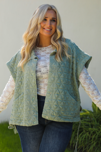 Close up of a woman model wearing a sage colored oversized vest paired with a white lace long sleeved top and dark denim outdoors.  The vest has functional pockets on either side, functional button closures in the front, and an oversized fit. 