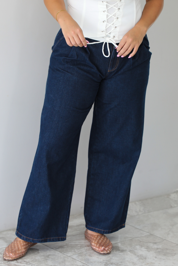 A person wearing dark blue wide-leg denim jeans with a white lace-up drawstring waist top, standing indoors against a white wall with white marble flooring. 