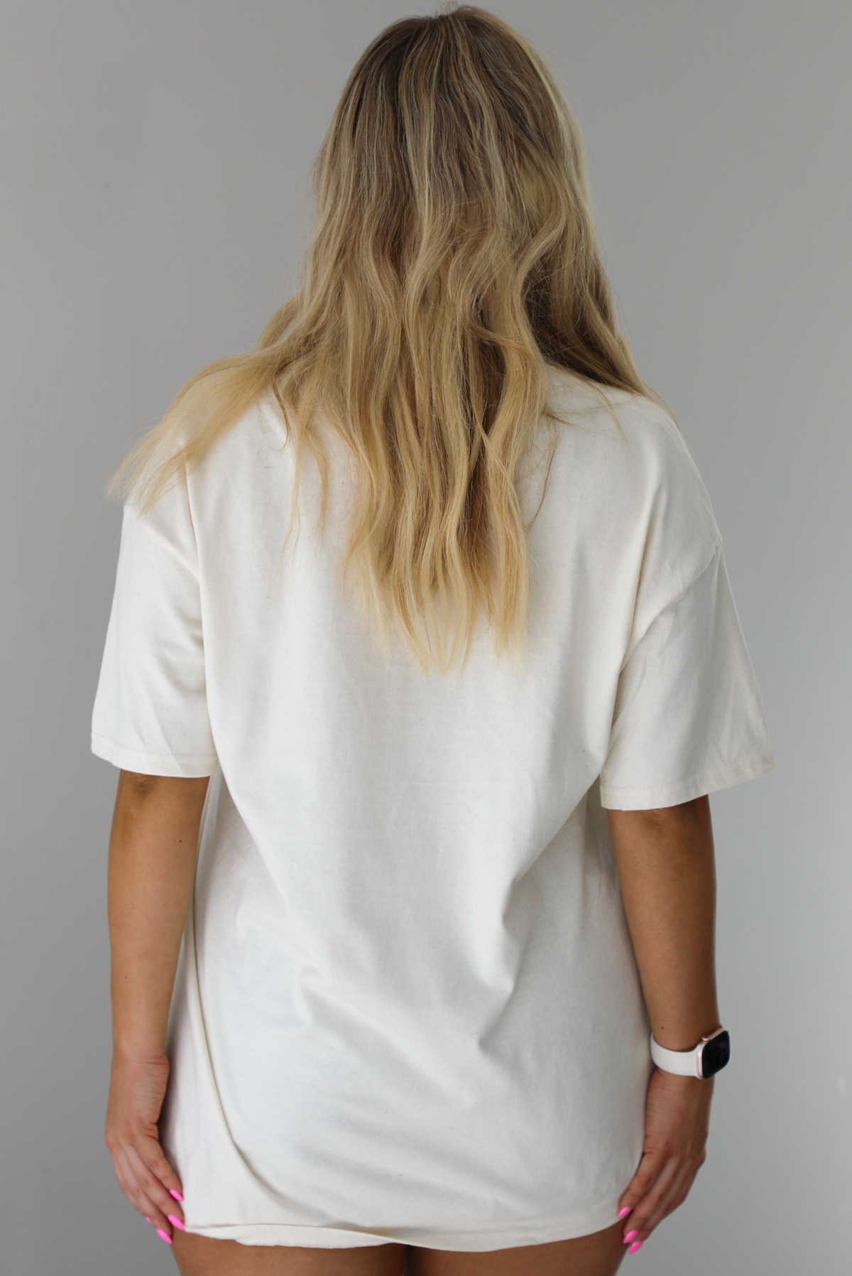 Woman wearing a white relaxed fit graphic tee that has wine, fruit, and flowers on it with the writing "Happiness Looks Good On You". The tee is untucked and paired with distressed denim shorts. The photo shows the back of the tee, which is plain. The photo is taken indoors against a white wall.