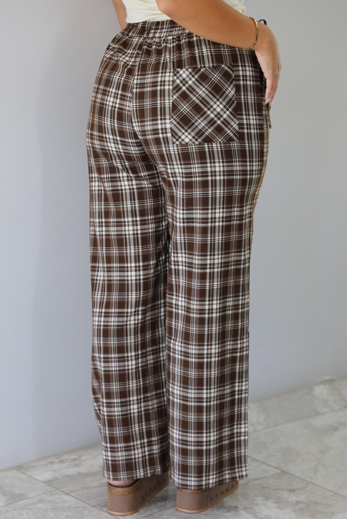 A person shown from the back wearing brown, white, and beige plaid wide-leg pants with a patch pocket, standing indoors against a white wall with white marble flooring. 