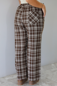 A person shown from the back wearing brown, white, and beige plaid wide-leg pants with a patch pocket, standing indoors against a white wall with white marble flooring. 