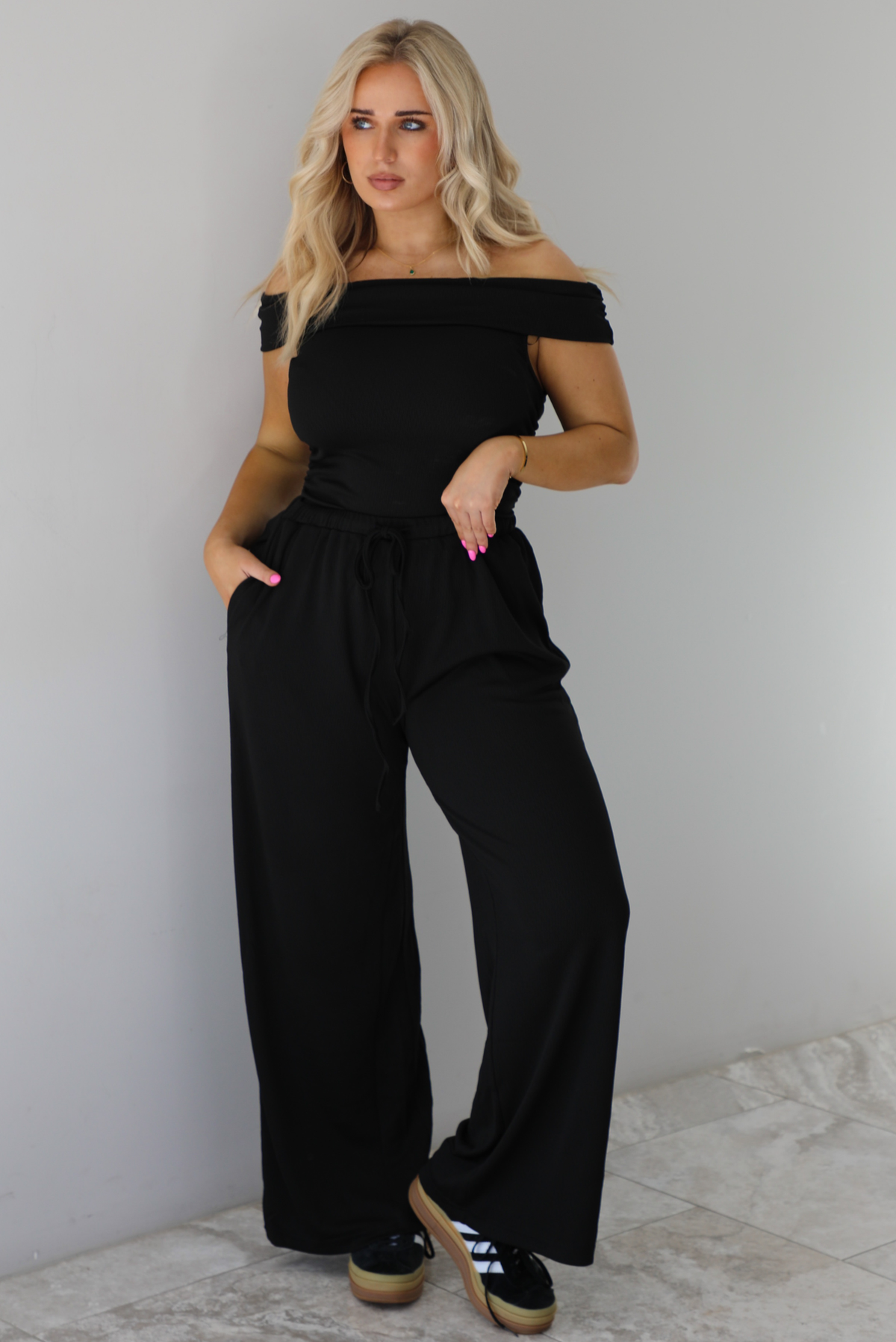 Woman wearing a two piece set featuring an off the shoulder top and high waisted pants. The photo is taken indoors against a white wall with white marble flooring. 