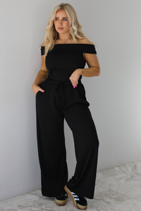 Woman wearing a two piece set featuring an off the shoulder top and high waisted pants. The photo is taken indoors against a white wall with white marble flooring. 