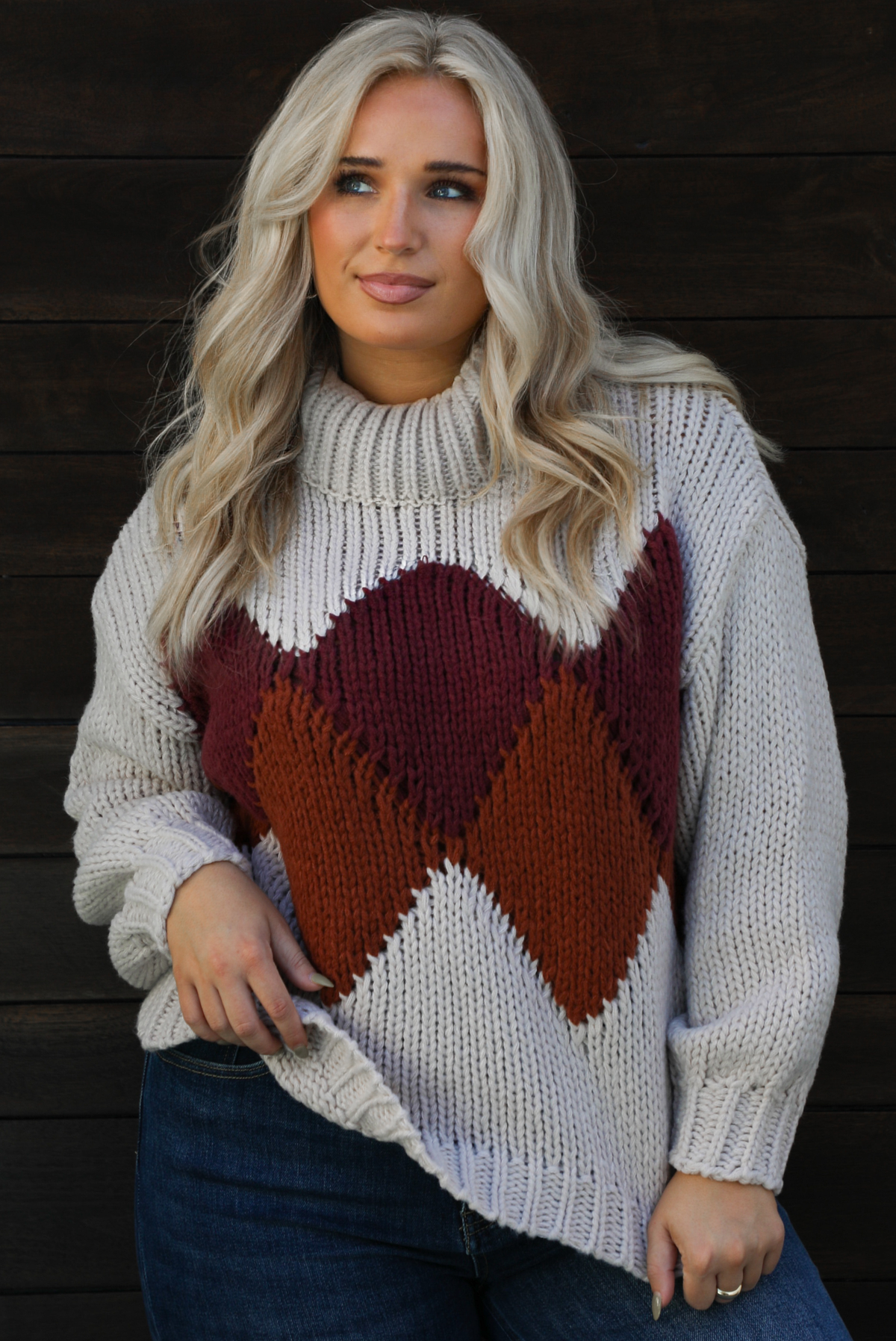 Close up of a woman model wearing a chunky knit turtle neck sweater with dark denim jeans outdoors in front of a dark wood door. 