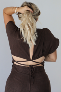 A woman shown from the back wearing a dark brown short-sleeve jumper, showing the tie closure in the back, with her hair in a high ponytail and bracelets on her wrist, against a plain light gray background.