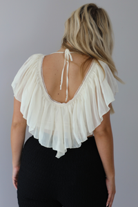 Woman wearing a cream ruffled cropped top with high waisted black, textured capri pants indoors against a white wall. The photo shows a closeup of the back of the top, which has a low cut back and a tie closure at the top. 