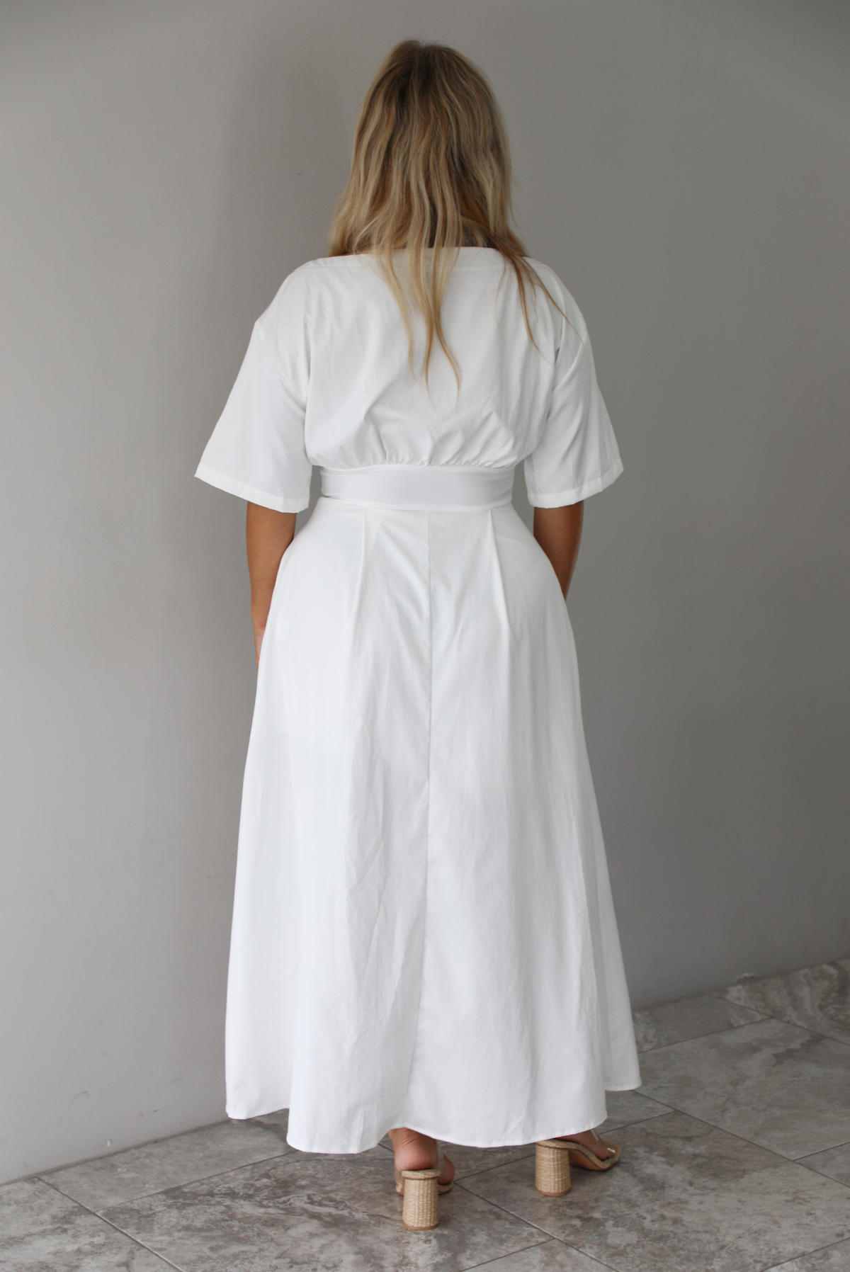 A woman shown from behind modeling a white midi dress with short puff sleeves, a wide boat neckline, and side pockets, standing indoors against a white wall with white marble flooring. 