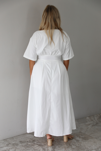 A woman shown from behind modeling a white midi dress with short puff sleeves, a wide boat neckline, and side pockets, standing indoors against a white wall with white marble flooring. 