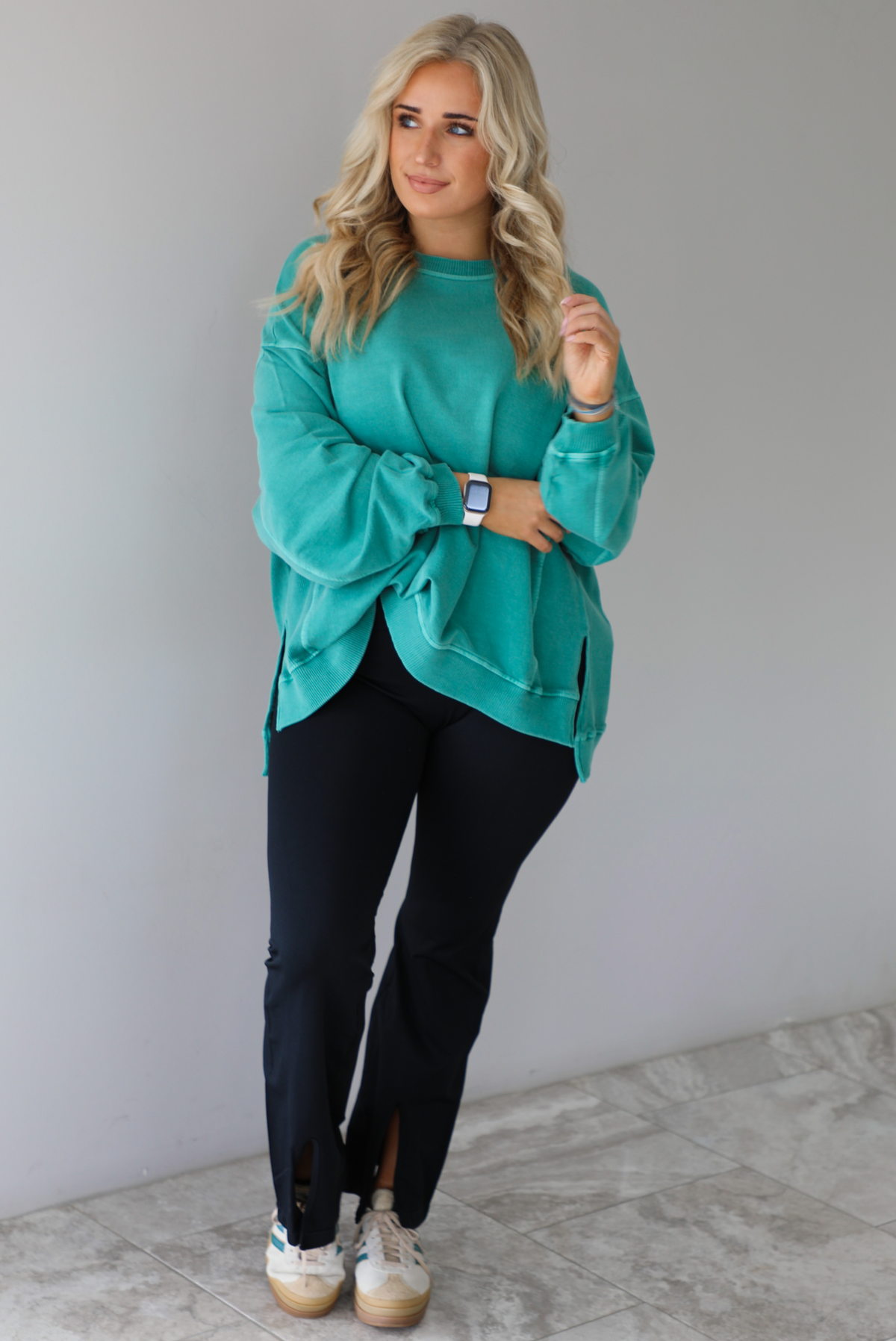 Woman wearing a teal oversized sweater with black leggings that have a slightly flared hem with split hem details in the front. The photo is taken in front of a white wall with white marble flooring. 