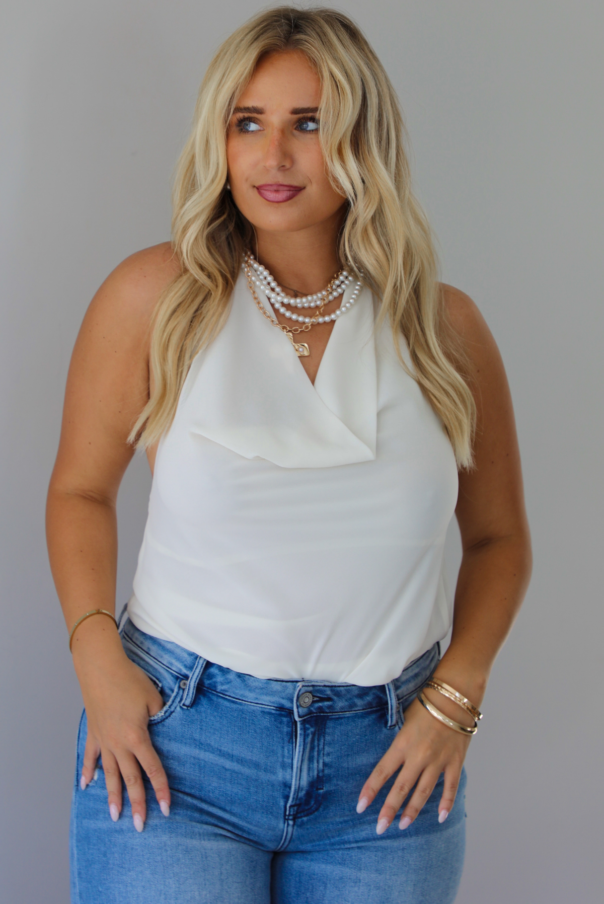 A woman modeling a cream sleeveless halter top and light-wash blue distressed flare jeans, standing indoors against a white wall.