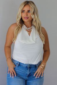 A woman modeling a cream sleeveless halter top and light-wash blue distressed flare jeans, standing indoors against a white wall.