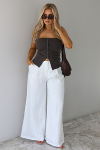 Woman wearing a strapless brown top with non-functional button closures in the front with  a split hem detail, and high waisted, flared hem white linen pants with a brown shoulder bag. The photo is taken indoors against a white wall with white marble flooring. 