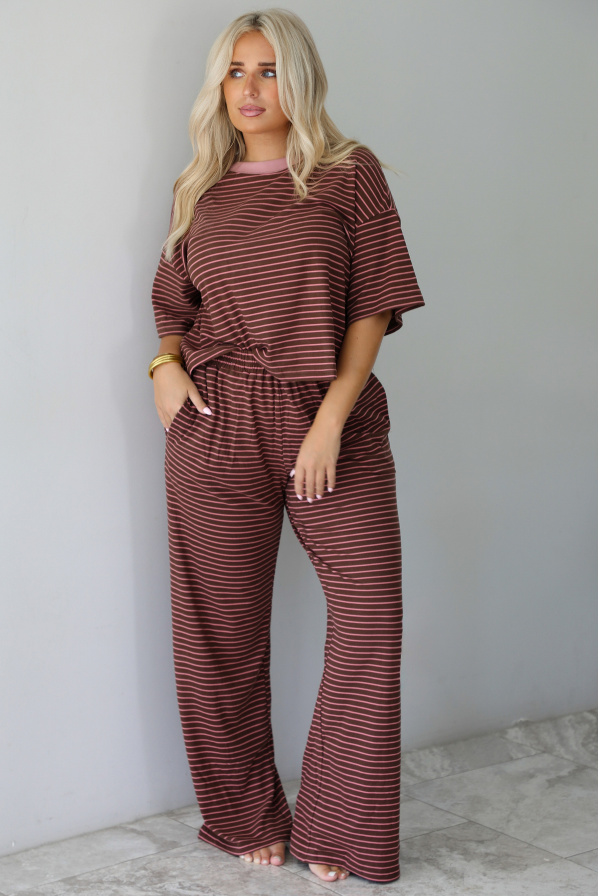 A woman wearing a brown and pink striped two piece set featuring a slightly cropped tee and high waisted pants, with the top slightly tucked into the pants, standing indoors against a white wall with white marble flooring. 