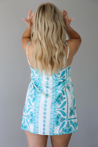 Woman wearing a teal and white patterned mini dress with thin, adjustable straps and a relaxed fit indoors against a white wall. The photo shows the back of the dress. 