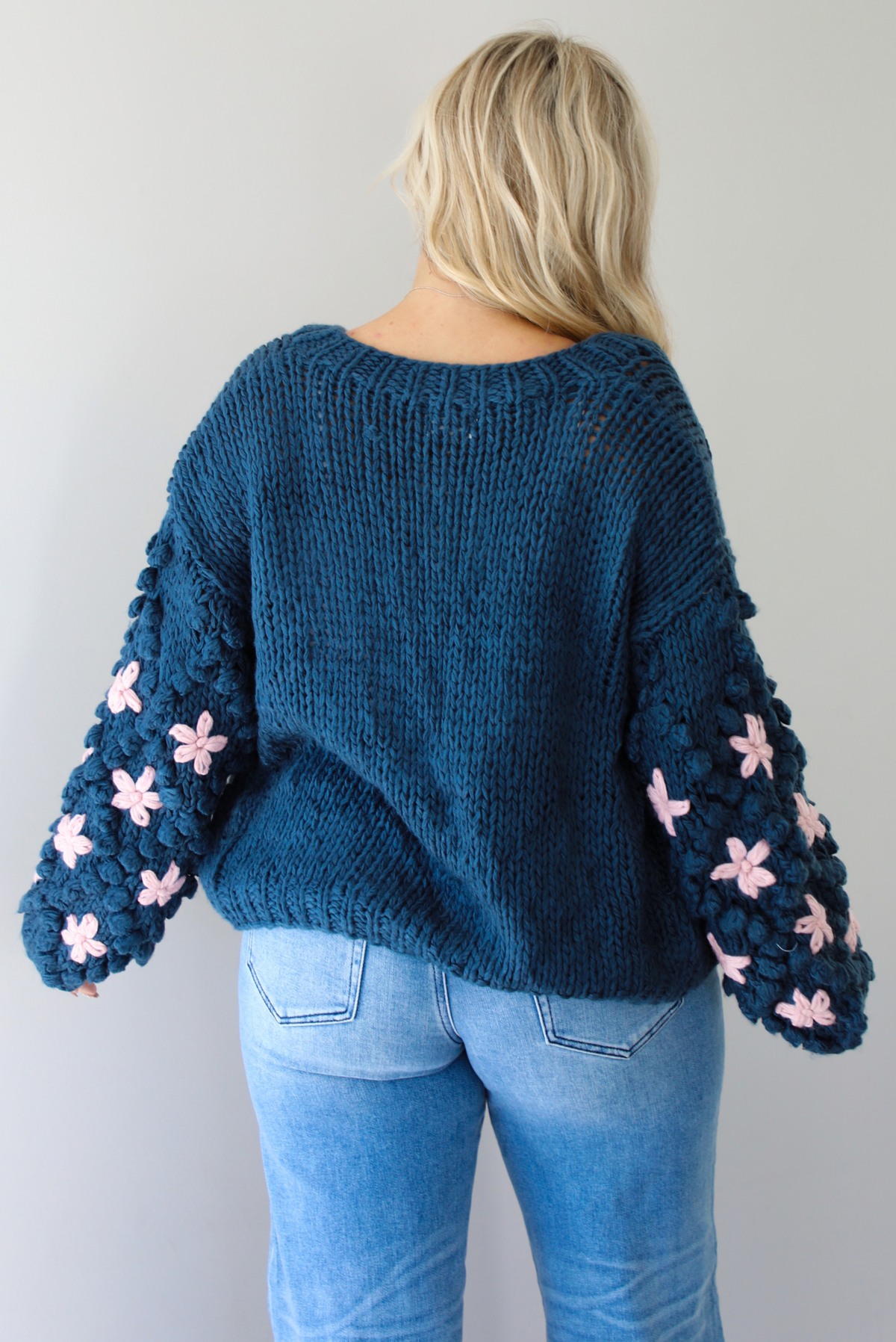 A woman shown from behind wearing a navy oversized cardigan with a pink embroidered floral design on the sleeves, paired with a white tank, mid-rise medium wash denim jeans, standing indoors against a white wall.