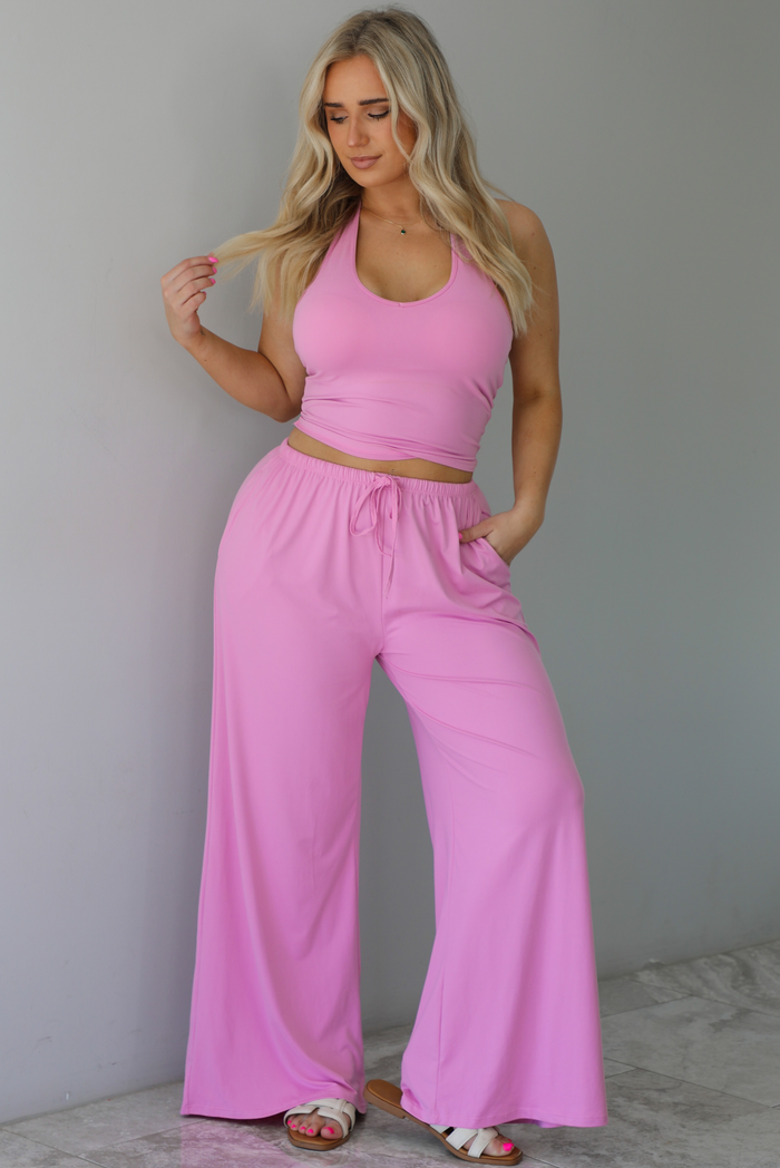 Woman wearing a two pieced pink set featuring a halter top and comfy pants with a flared hem detail. The photo is taken indoors against a white wall with white marble flooring. 