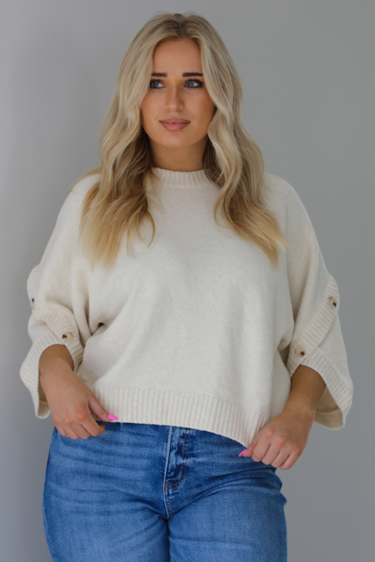 Woman wearing a cream colored quarter sleeve sweater that has ribbed details at the neckline, on the sleeves, and on the hem, with non-functional button closures on the sleeves. The sweater has a relaxed fit and is untucked from the denim to show the full length. The photo is taken indoors against a white wall.