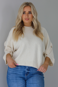 Woman wearing a cream colored quarter sleeve sweater that has ribbed details at the neckline, on the sleeves, and on the hem, with non-functional button closures on the sleeves. The sweater has a relaxed fit and is untucked from the denim to show the full length. The photo is taken indoors against a white wall.
