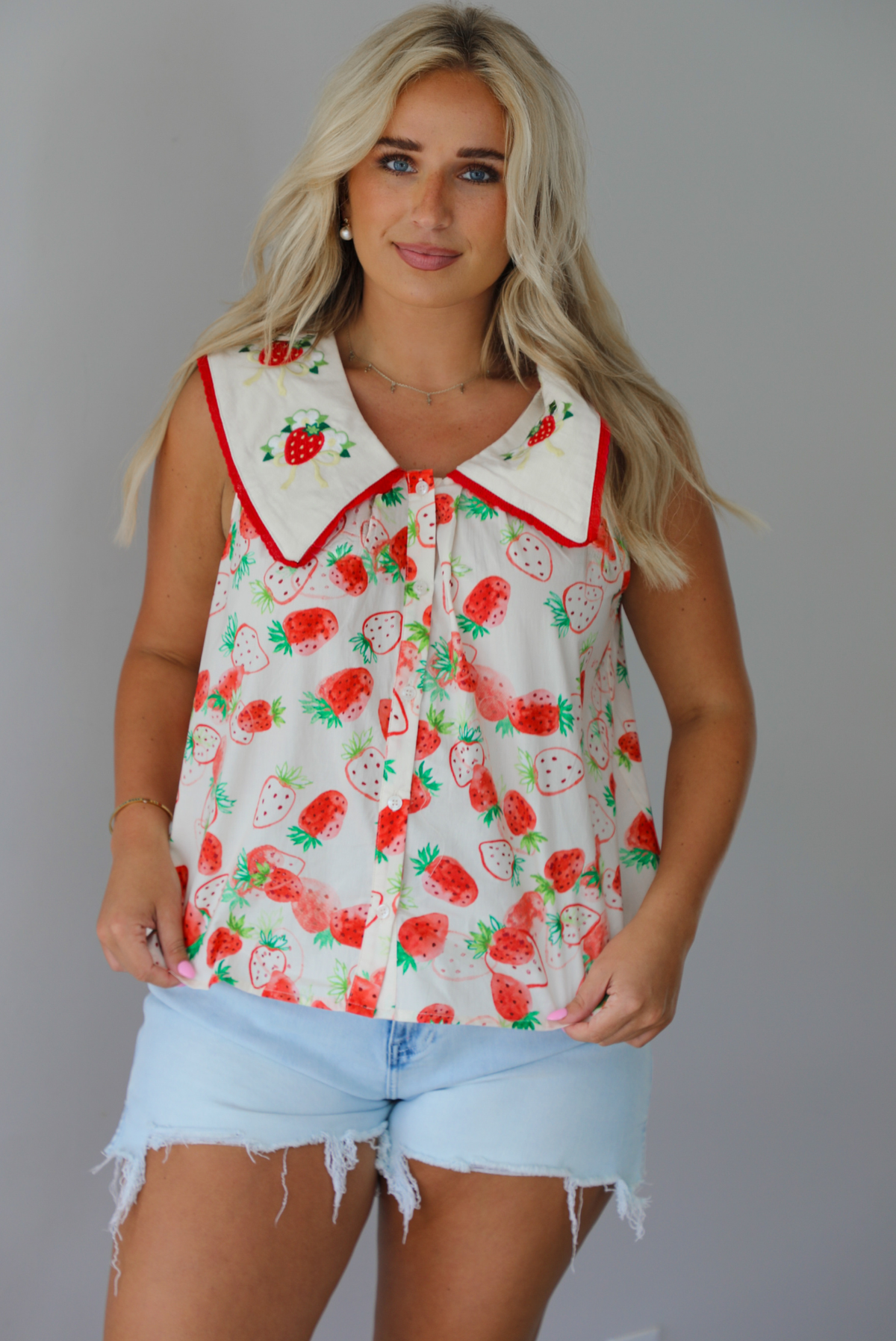 Woman wearing a cream and red strawberry patterned top with a collared detail and functional button closures in the front with light wash denim shorts indoors against a white wall. The photo shows the top untucked to show its full length. 
