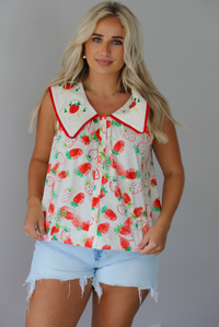 Woman wearing a cream and red strawberry patterned top with a collared detail and functional button closures in the front with light wash denim shorts indoors against a white wall. The photo shows the top untucked to show its full length. 