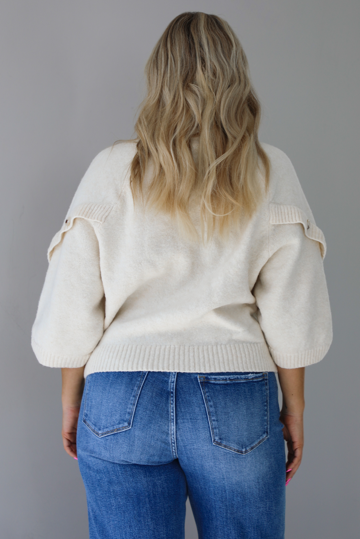 Woman wearing a cream colored quarter sleeve sweater that has ribbed details at the neckline, on the sleeves, and on the hem, with non-functional button closures on the sleeves. The sweater has a relaxed fit and is untucked from the denim to show the full length in the back. The photo is taken indoors against a white wall.