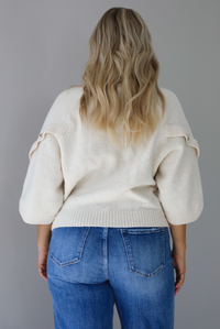 Woman wearing a cream colored quarter sleeve sweater that has ribbed details at the neckline, on the sleeves, and on the hem, with non-functional button closures on the sleeves. The sweater has a relaxed fit and is untucked from the denim to show the full length in the back. The photo is taken indoors against a white wall.