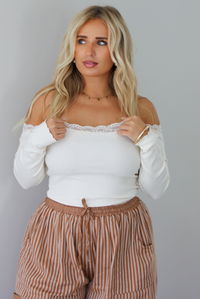 A closeup of a woman wearing a cream off the shoulder long sleeved top tucked into a pair of camel and cream striped pants, standing indoors against a white wall. 