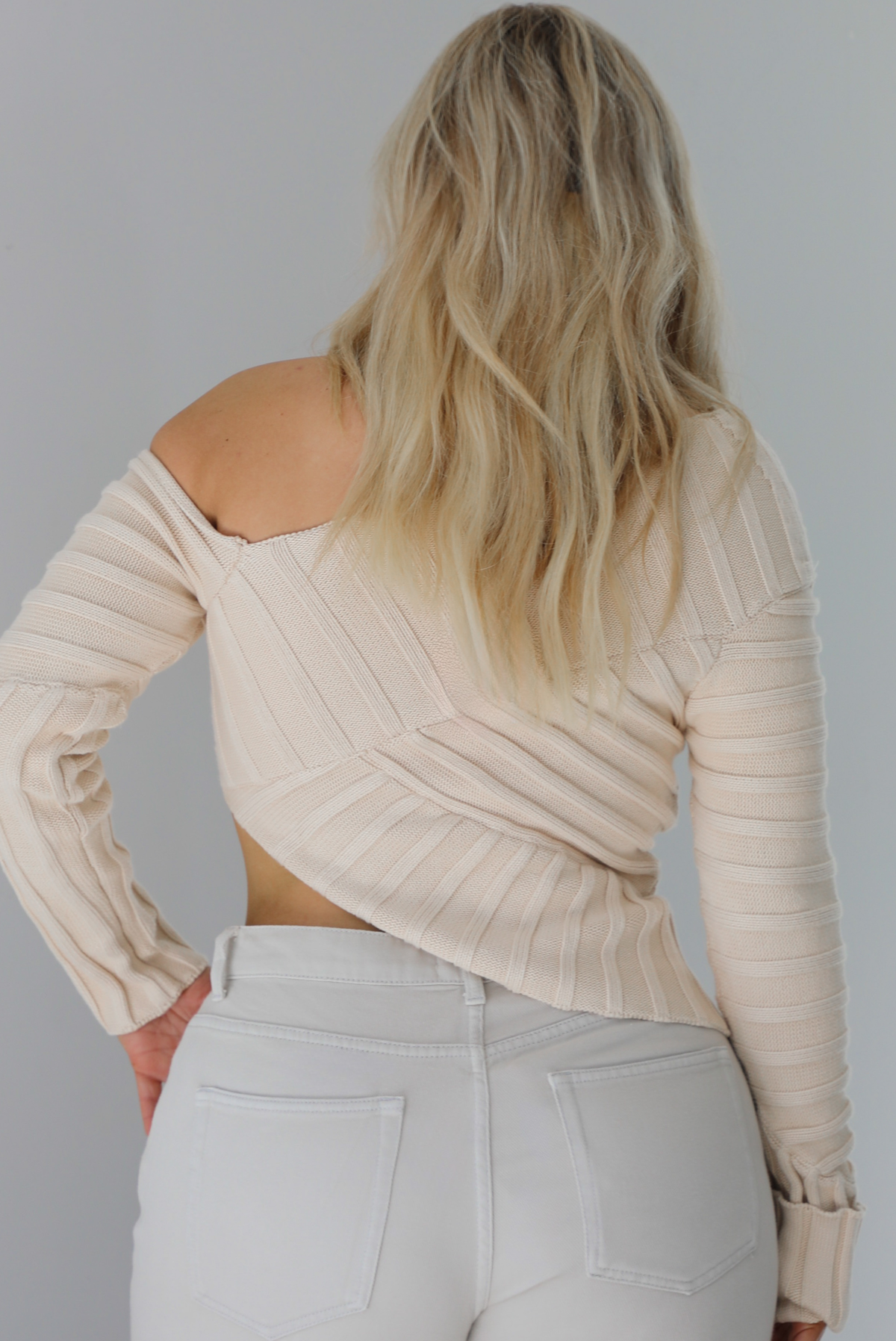 Woman wearing a cream colored ribbed textured knit top that has an asymmetrical hem with long sleeves in which one side is worn off the shoulder. The sweater is paired with sand colored high waisted, cuffed hem denim. The photo shows a closeup of the back of the knit top. The photo is taken indoors against a white wall.