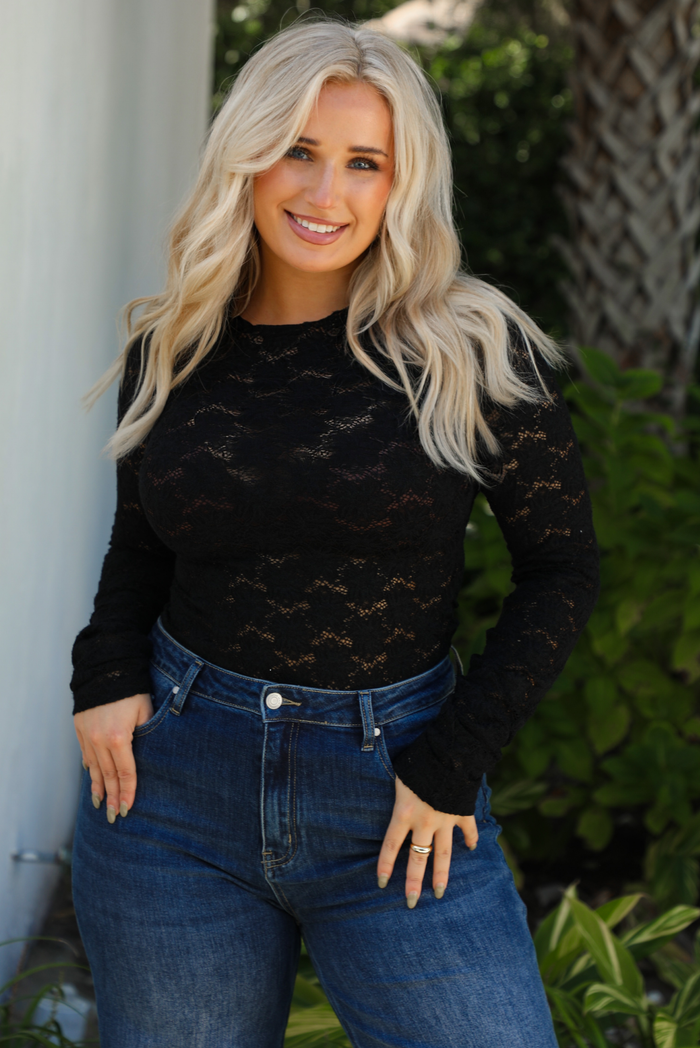 Close up of model wearing black sheer lace top with a floral pattern with dark denim jeans outdoors.