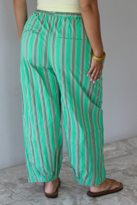 A woman wearing a pale yellow tank and mid-rise pants that have a green, brown, lime green, and tan striped pattern, which have an elastic waistline, functional pockets on either side, and cinched details at the ankles, standing indoors against a white wall with white marble flooring.  The photo is close up and focused on the back of the pants. 