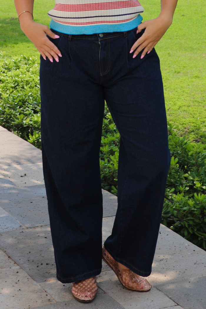 A person wearing dark navy wide-leg jeans, standing outdoors on a stone path with green grass and bushes in the background.