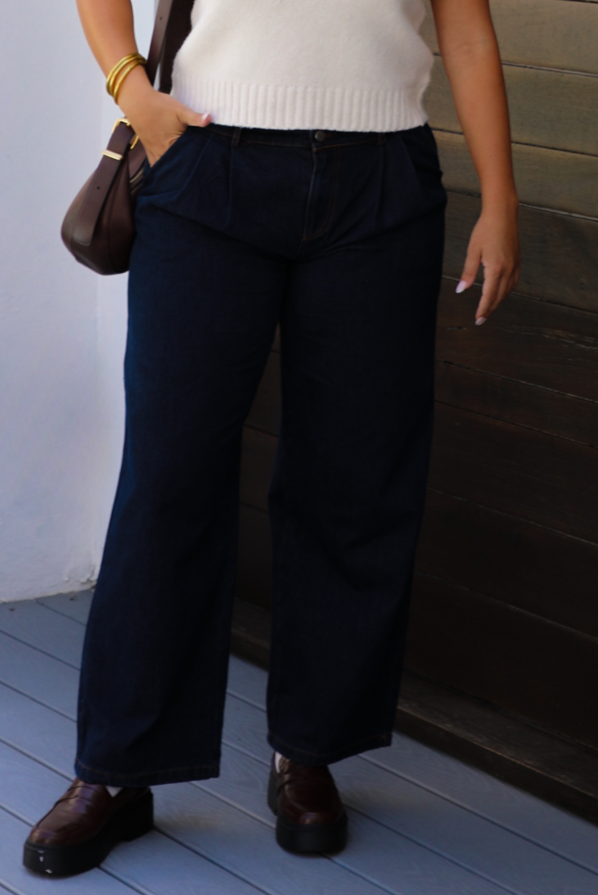 A woman wearing dark denim wide-leg jeans, a cream knit top, and brown leather shoes, standing outdoors against a dark wooden wall.