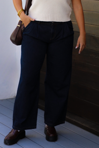 A woman wearing dark denim wide-leg jeans, a cream knit top, and brown leather shoes, standing outdoors against a dark wooden wall.