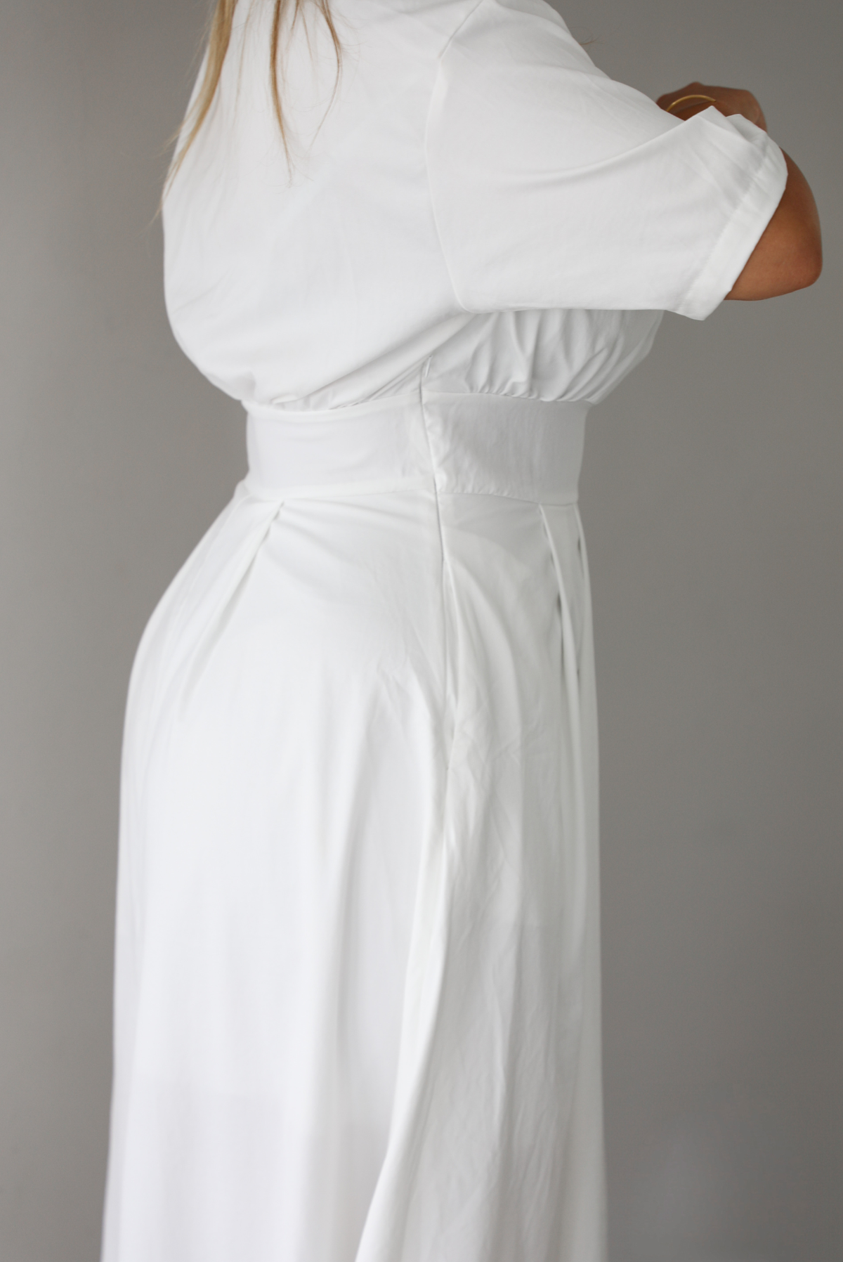 A close up of a woman modeling a white midi dress with short puff sleeves, a wide boat neckline, and side pockets, standing indoors against a white wall, focusing on the zipper closure on the side. 