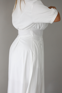 A close up of a woman modeling a white midi dress with short puff sleeves, a wide boat neckline, and side pockets, standing indoors against a white wall, focusing on the zipper closure on the side. 