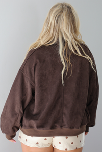 A woman shown from behind wearing a brown suede jacket, paired with a white tank and cream colored mini skort with a brown polka dot print pattern throughout, standing indoors against a white wall.