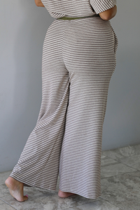A person shown from behind wearing loose, wide-leg striped pants with a muted green drawstring waistband and a matching striped crop top, standing barefoot against a plain white wall with white marble flooring. 