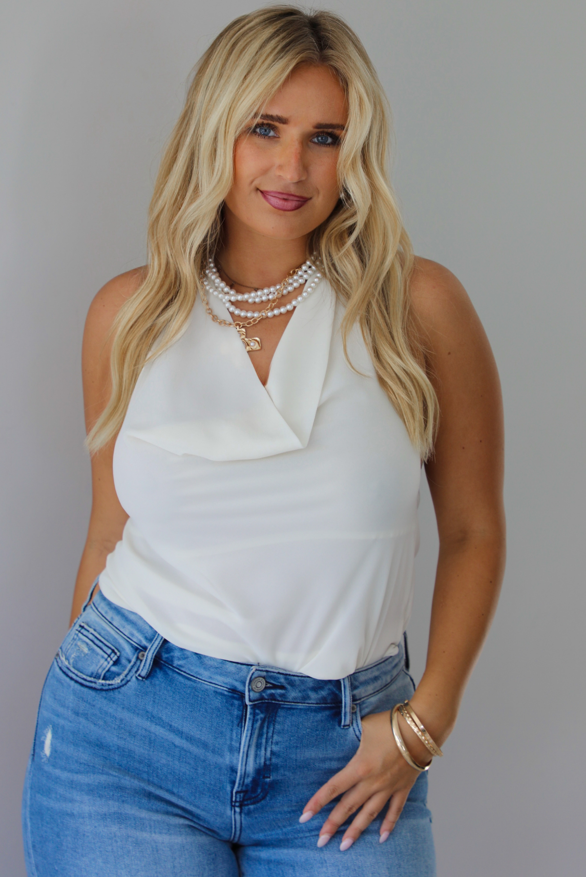 A woman modeling a cream sleeveless halter top and light-wash blue distressed flare jeans, standing indoors against a white wall.