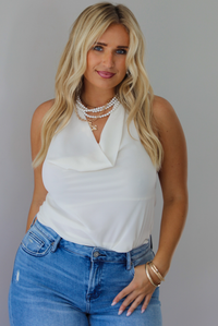 A woman modeling a cream sleeveless halter top and light-wash blue distressed flare jeans, standing indoors against a white wall.