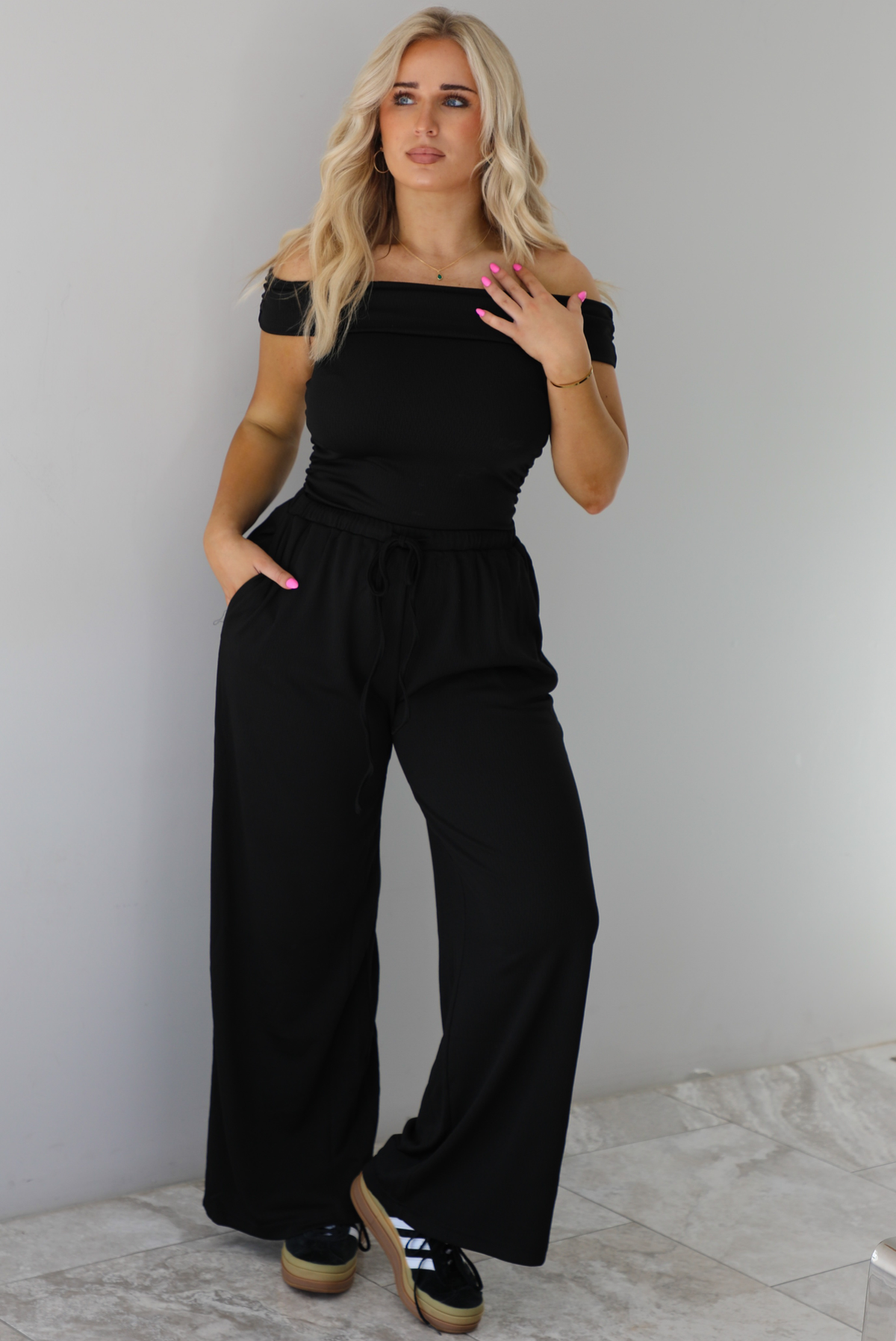 Woman wearing a two piece set featuring an off the shoulder top and high waisted pants. The photo is taken indoors against a white wall with white marble flooring. 