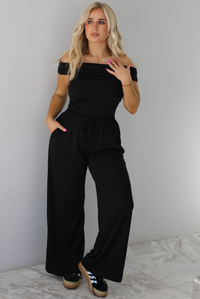 Woman wearing a two piece set featuring an off the shoulder top and high waisted pants. The photo is taken indoors against a white wall with white marble flooring. 