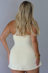 A woman shown from behind wearing a pale yellow ribbed athleisure dress with a knotted waist skirt detail, standing indoors against a white wall.