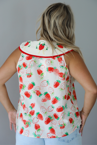 Woman wearing a cream and red strawberry patterned top with a collared detail and functional button closures in the front with light wash denim shorts indoors against a white wall. The photo shows the back of the top. 