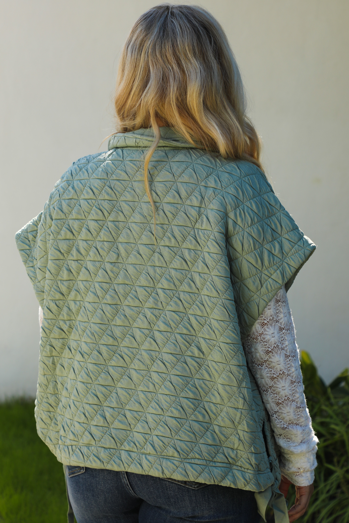 Close up of the back of a woman model wearing a sage colored oversized vest paired with a white lace long sleeved top and dark denim outdoors.  The vest has functional pockets on either side, functional button closures in the front, and an oversized fit. 