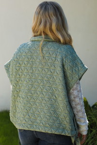 Close up of the back of a woman model wearing a sage colored oversized vest paired with a white lace long sleeved top and dark denim outdoors.  The vest has functional pockets on either side, functional button closures in the front, and an oversized fit. 